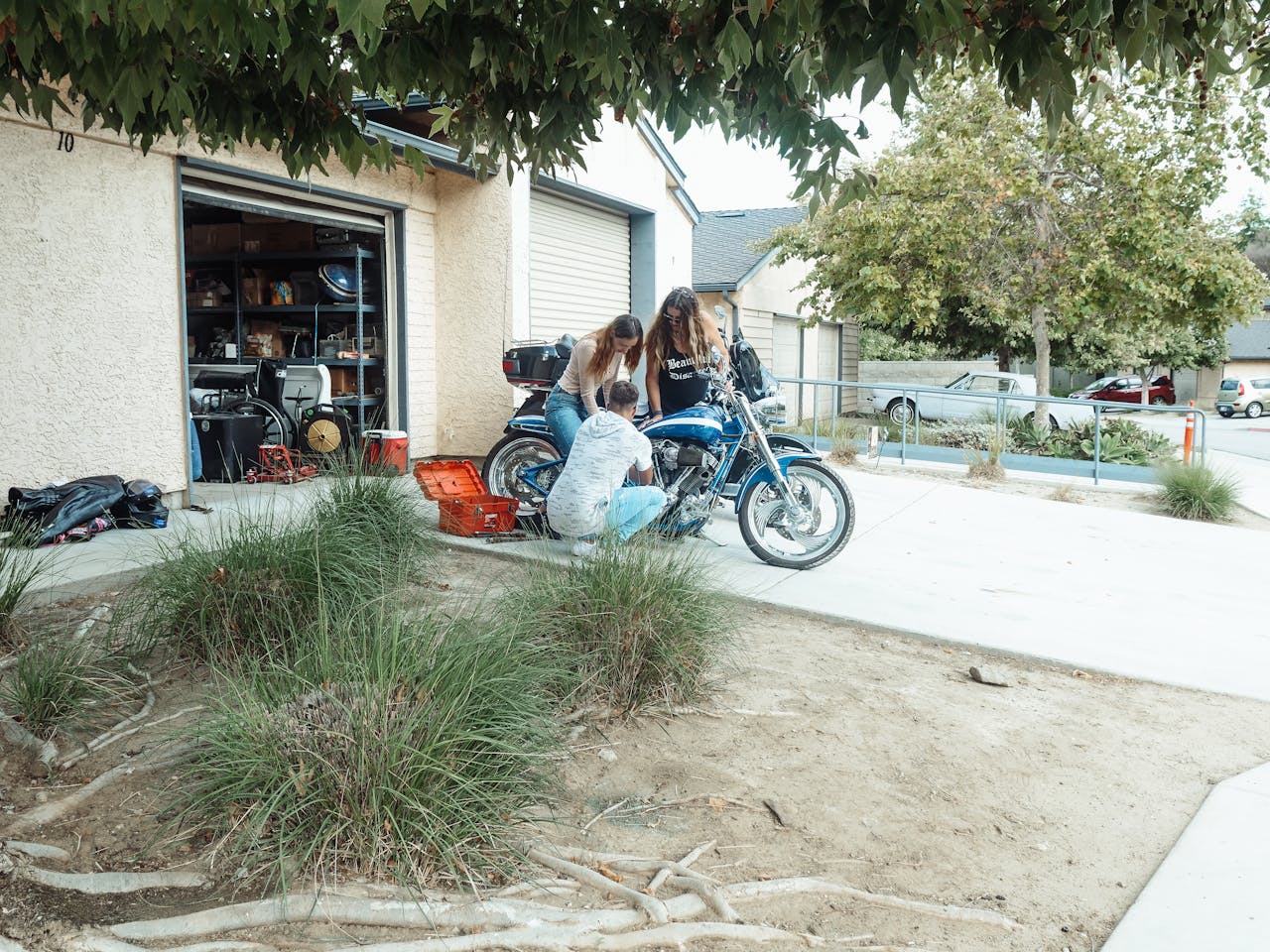 Two people repairing a motorcycle outside a suburban garage. Tools and greenery around.